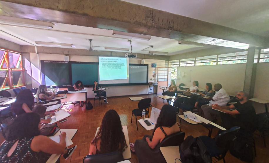 Imagem de uma das oficinas do evento que aconteceu em uma sala de aula. As pessoas formaram um círculo com as carteiras e estão atentas ao que fala uma das participantes.