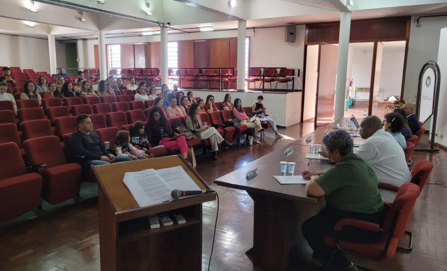 Imagem que mostra a mesma mesa de abertura do evento e também a plateia, formada por diversas servidoras e servidores da Defensoria.