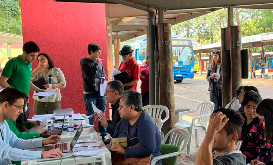 Imagem que mostra algumas pessoas aguardado atendimento e outras sendo atendias no mutirão realizado no TTU de Foz do Iguaçu vista de outro ângulo. Ao fundo, um ônibus azul está parado no terminal.