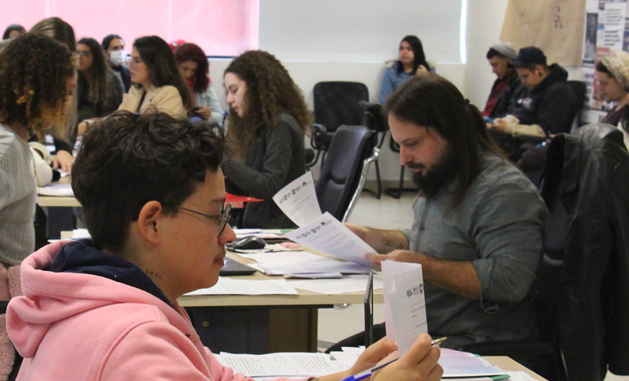 Imagem que mostra uma pessoa sendo atendida em Curitiba, ela está segurando um papel e olhando para ele. Ao fundo, outras pessoas aguardam atendimento.