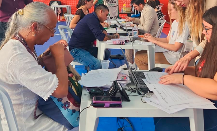 Imagem que mostra uma mulher sendo atendida por profissionais da DPE-PR no mutirão em Paranaguá. Elas estão em uma mesa com cadeiras plásticas, brancas, em um ginásio. Ao fundo, mais mesas de atendimento e pessoas sendo atendidas.