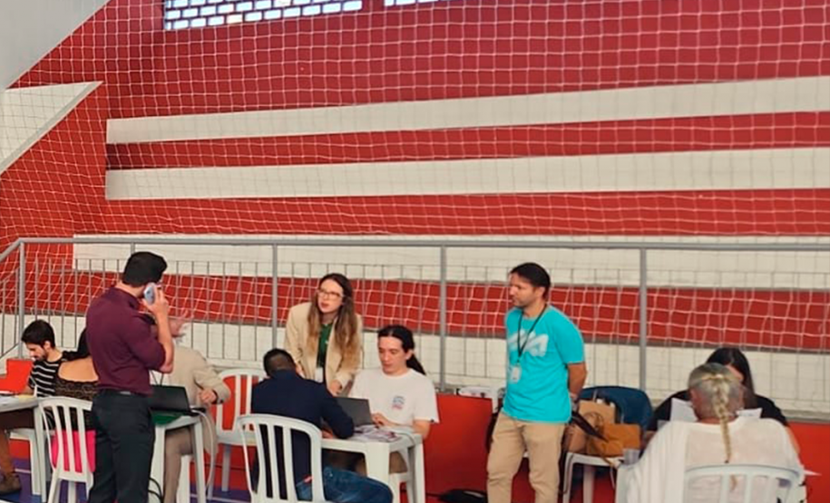 Imagem que mostra o atendimento no mutirão em Paranaguá, com três mesas com cadeiras plásticas, brancas, em um ginásio. Nas três estão pessoas sendo atendidas.