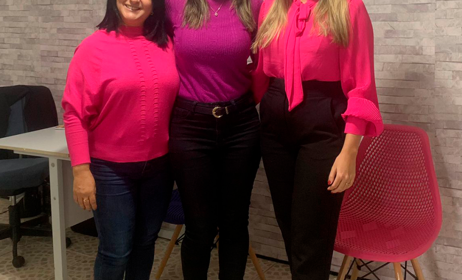 Na foto estão pousando as secretárias de atendimento Daiane Cristiane Pereira dos Santos e Dioneia dos Santos Modesto, e a defensora pública de Paranaguá Ingrid Lima Vieira, nesta ordem da esquerda para a direitas. As três estão sorrindo, com roupas cor de rosa e roxa.