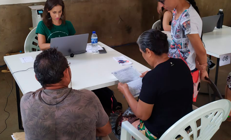 Imagem que mostra um atendimento no mutirão. Três pessoas estão sendo atendidas por uma mulher, a defensora pública Aline Bastos, que está olhando para um notebook. Duas pessoas que estão sendo atendidas analisam um documento.