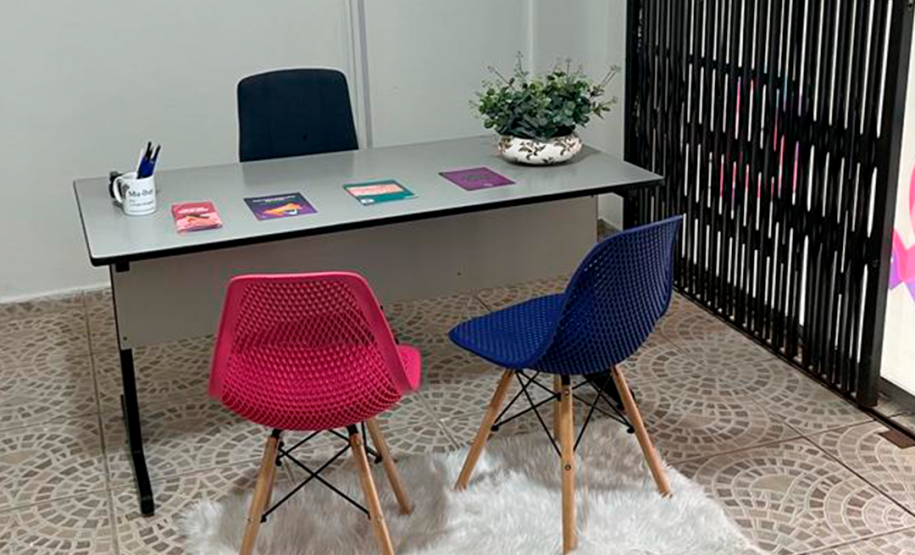 Fotografia da sala de atendimento da Defensoria Pública do Estado do Paraná no Ponto de Atendimento à Mulher. A foto mostra uma mesa com a cadeira da defensora pública de um lado, e do outro duas cadeiras, uma roxa e outra rosa, onde a usuária poderá sentar.
