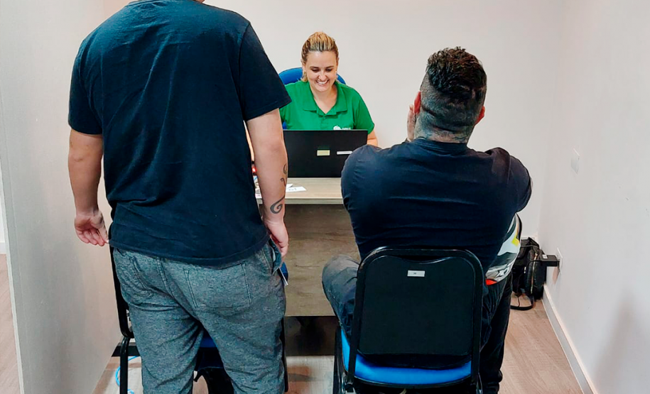 Imagem da assessora Bruna Abdalla atendendo dois usuários da DPE-PR, um deles está de pé e o outro está sentado. Ela está sentada de frente para eles e sorri.