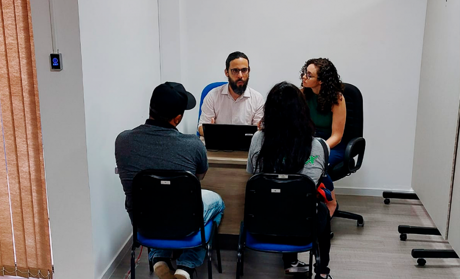 Imagem que mostra o defensor Fernando Redede e a psicóloga Lethicia Gaidarji Silva atendendo a um casal de usuários da DPE-PR, todos estão sentados em volta de uma mesa.