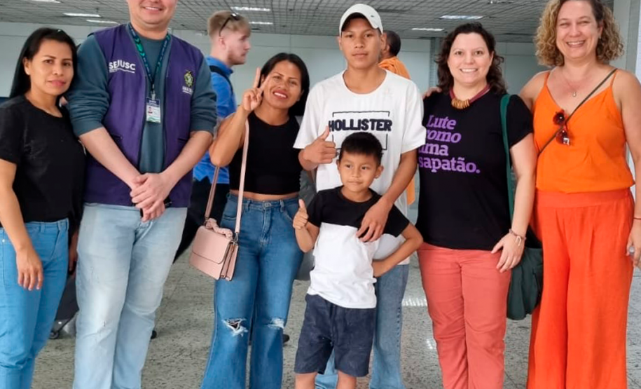 Imagem da família mencionada na matéria, a foto conta com a família acompanhada da equipe que auxiliou o reencontro, eles estão posando para a foto no aeroporto.