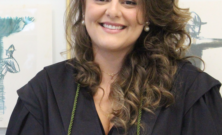 Imagem da nova defensora pública, Gabriela Vizel Gomes. Ela olha para frente, sorrindo. Está com os cabelos soltos, que são cacheados, um pouco abaixo do ombro. Ela usa uma beca preta, com um cordão verde.