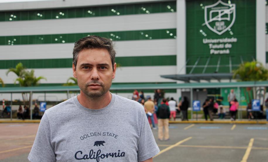 Imagem que mostra o candidato João Carlos de Carvalho em frente ao local de prova, um dos prédios da Universidade Tuiuti, em Curitiba. Ele veste uma camiseta cinza e olha para a frente.