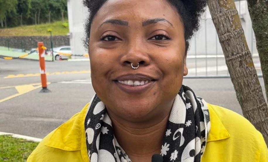 Imagem que mostra a candidata Leoni Rita Vitória sentada em frente ao local de prova, em Curitiba. Ela veste uma camiseta amarela e um lenço nas cores preta e branca.