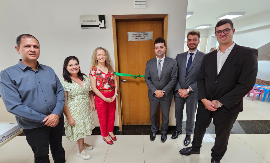 Imagem que mostra a inauguração da sala da DPE-PR em Cruzeiro do Oeste. Estão presentes a juíza diretora do Fórum, a promotora de Justiça Wilza Lacerda, o coordenador regional do Deppen, o defensor público-geral, o defensor público Pedro Bruzzi, e o 2º subdefensor-geral do estado. Todos estão ao redor da porta da sala, onde há uma placa que diz “Defensoria Pública do Estado do Paraná”. Cruzando a porta de um lado a outro há uma fita de cetim de cor verde, com um laço ao meio. Todos sorriem.