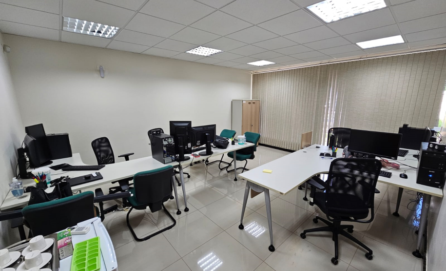 Imagem que mostra o interior da sala da DPE-PR em Cruzeiro do Oeste, onde é possível ver duas grandes mesas com computadores e cadeiras para pessoas que serão atendidas. Ao fundo, há uma grande janela, que está ocultada por uma persiana.