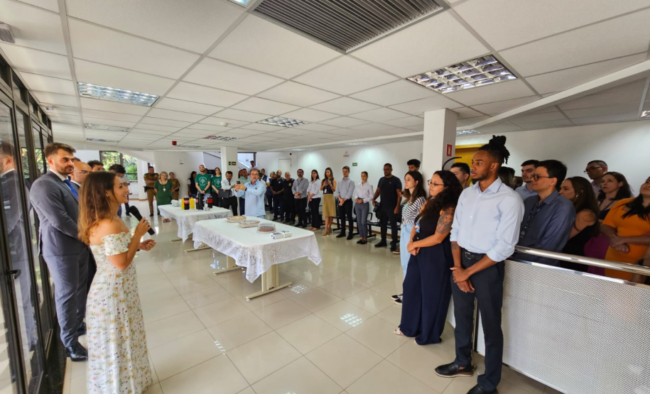 Imagem que mostra a inauguração da sala da DPE-PR no Fórum de Cambé, onde a defensora pública Aline Bastos discursa para várias pessoas que assistem a cerimônia. Todos estão em pé, em um hall amplo e bem iluminado, dentro do Fórum. Há também duas mesas no centro, com comidas e bebidas.