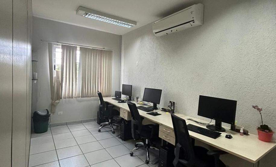 Imagem que mostra a sala de atendimento da DPE-PR em Jandaia do Sul, onde há uma mesa comprida, com 3 computadores com teclados e mouses, e 3 cadeiras. Na parede, acima da mesa, há um aparelho de ar condicionado. Ao fundo, há uma janela com uma cortina entreaberta.