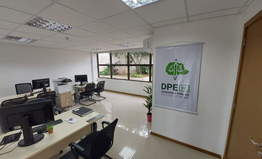 Imagem que mostra outra sala da DPE-PR no Fórum de Cambé, onde há um banner com o logo da Defensoria na parede ao lado da porta e três mesas com computadores e cadeiras para pessoas serem atendidas. Ao fundo é possível ver uma grande janela que mostra uma parte externa do Fórum, onde há algumas plantas em um canteiro.