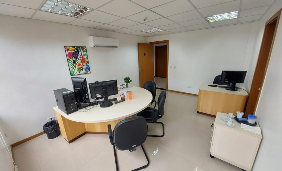 Imagem que mostra uma das salas da DPE-PR no Fórum de Cambé, onde há uma mesinha com um computador e, ao lado, mais ao centro da sala, uma mesa maior, redonda, com um computador com dois monitores e cadeiras ao redor, para receber pessoas que serão atendidas. Na parede, acima dessa mesa maior, há um quadro com desenhos de plantas e pássaros e um aparelho de ar condicionado.