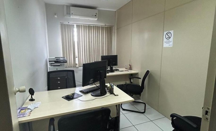 Imagem que mostra a sala de atendimento da DPE-PR em Jandaia do Sul, onde há duas mesas, com computadores com teclados e mouses, e cadeiras para quem usa o computador e para pessoas que estejam sendo atendidas. Ao fundo, há uma janela com uma cortina entreaberta. Acima, um aparelho de ar condicionado e, ao lado da janela, uma impressora multifuncional.