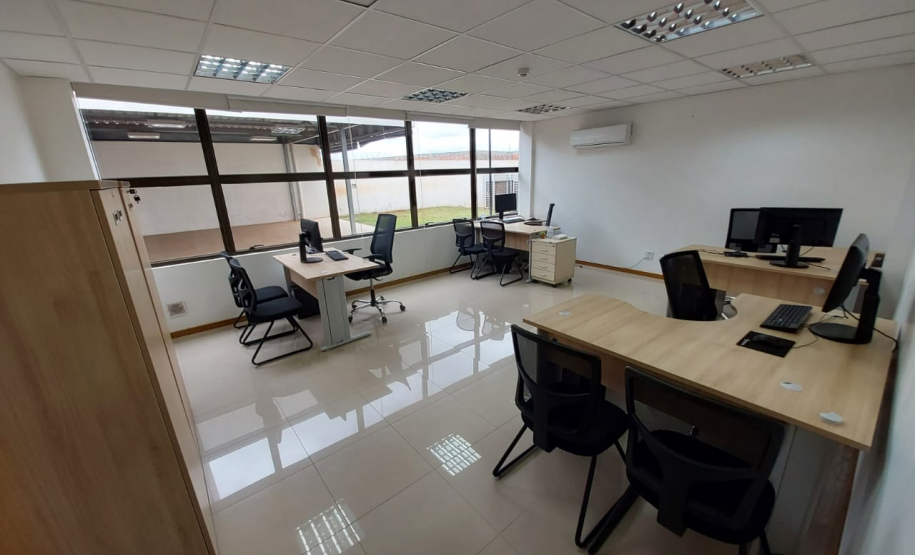 Imagem que mostra outra sala da DPE-PR no Fórum de Cambé, onde há um armário e quatro mesas com computadores e cadeiras para pessoas serem atendidas. Ao fundo é possível ver uma grande janela que mostra uma parte externa do Fórum.