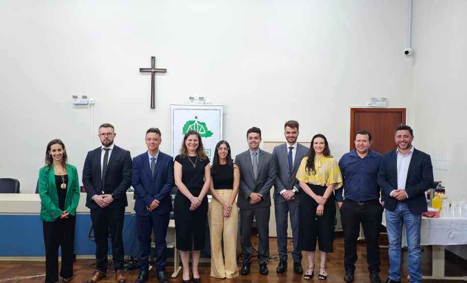Imagem que mostra representantes da Defensoria e autoridades convidadas na inauguração da sala da DPE-PR em Jandaia. As pessoas, 4 mulheres e 6 homens, estão lado a lado e sorriem. Elas estão em uma sala ampla do Fórum, onde foi realizada a solenidade. Atrás delas é possível ver um banner com o logo da Defensoria e um crucifixo.