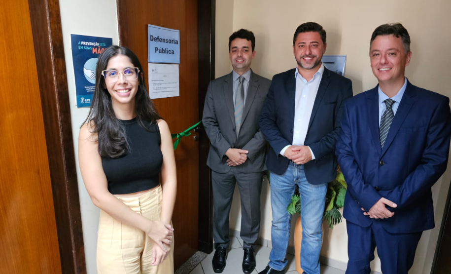 Imagem que mostra a defensora pública Maria Luiza Valverde, o defensor público-geral, André Giamberardino, e o prefeito de Jandaia do Sul, Lauro Junior, diante da porta da sala da Defensoria no Fórum de Jandaia. Na porta, há uma placa que diz “Defensoria Pública” e cruzando a porta de um lado a outro há uma fita de cetim de cor verde, com um laço ao meio. Todos sorriem.