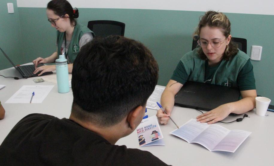 Imagem de uma foto com um ângulo fechado de um atendimento, onde uma servidora pública apresenta o trifolder sobre o mutirão Meu Nome, Meu Direito para um usuário.