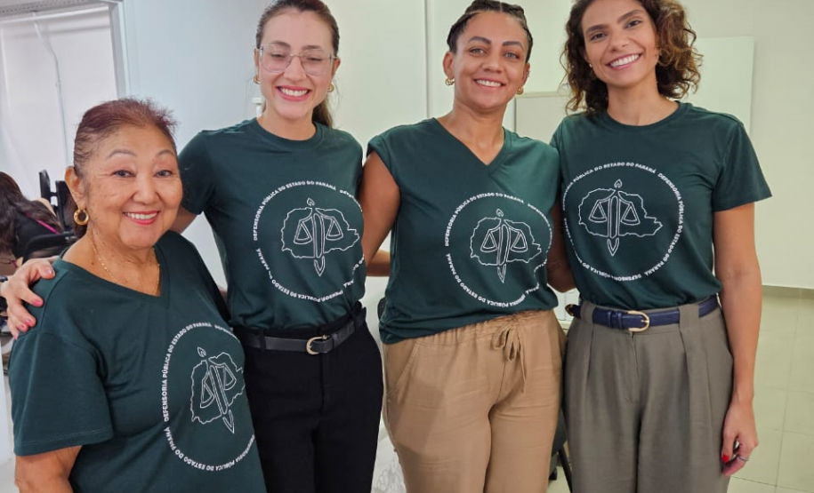 Imagem de uma foto espontânea de servidoras públicas, todas usando uma camiseta verde da Defensória Pública do Estado do Paraná. No fundo da imagem, dá para ver uma bandeira LGBTQIA+ pendura do teto.