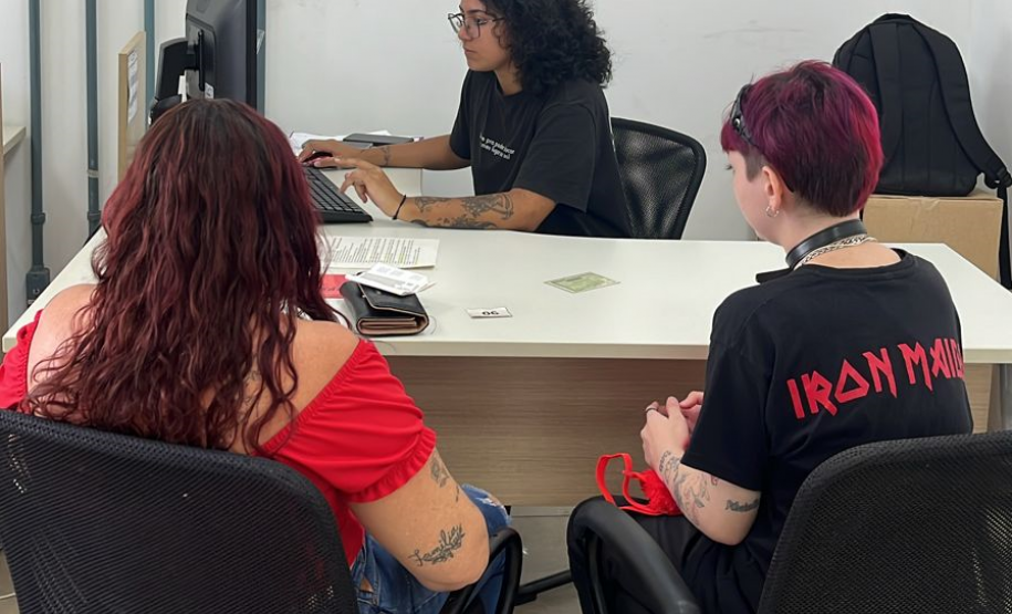 Imagem que mostra duas pessoas sendo atendidas no mutirão em Ponta Grossa. Elas estão de costas e entre elas, é possível ver a pessoa que está fazendo o atendimento, sentada atrás da mesa e olhando para a tela do computador.