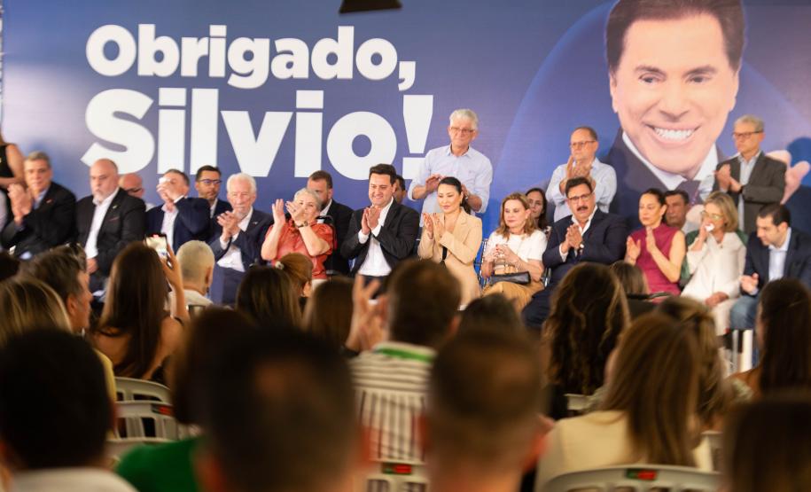 Imagem colorida horizontal. No primeiro plano se vê o público observando autoridades no lançamento do projeto de construção do Centro de Reabilitação Silvio Santos. Ao fundo cartaz com a imagem do apresentador onde se lê: Obrigado, Silvio.