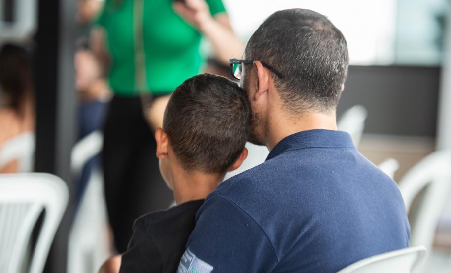 Imagem horizontal colorida em que se vê um pai com o filho no colo de costas enquanto aguarda o atendimento.