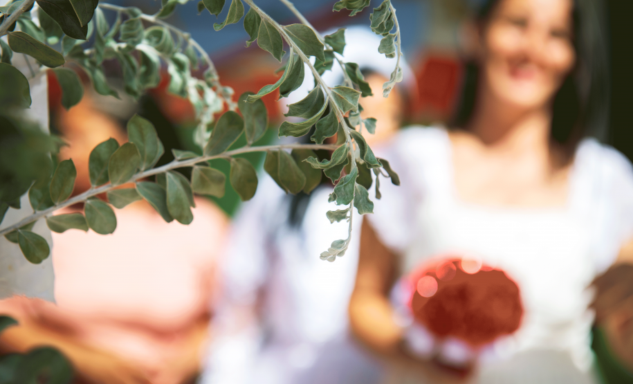 Imagem que mostra em primeiro plano um ramo de planta, com folhas verdes. Ao fundo, uma das noivas sorri e segura um pequeno buquê de flores vermelhas. Essa parte da imagem está borrada, para preservar a identidade dela.