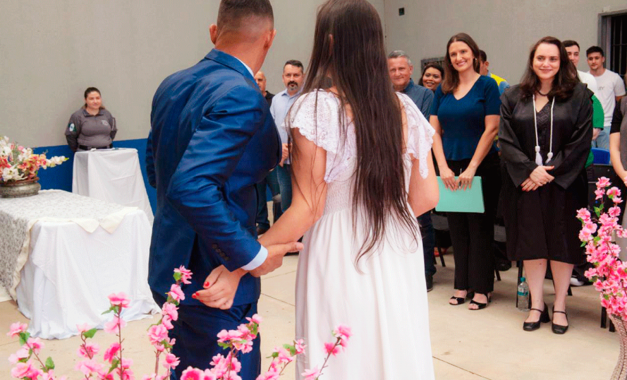 Imagem que mostra um dos casais, de costas. Ele usa um terno azul e ela um vestido branco. Ela está com o cabelo solto, liso, que vai até o meio das costas. Ao fundo, as autoridades presentes assistem a cena e sorriem. Arranjos com flores rosas em arranjos compõem a cena.