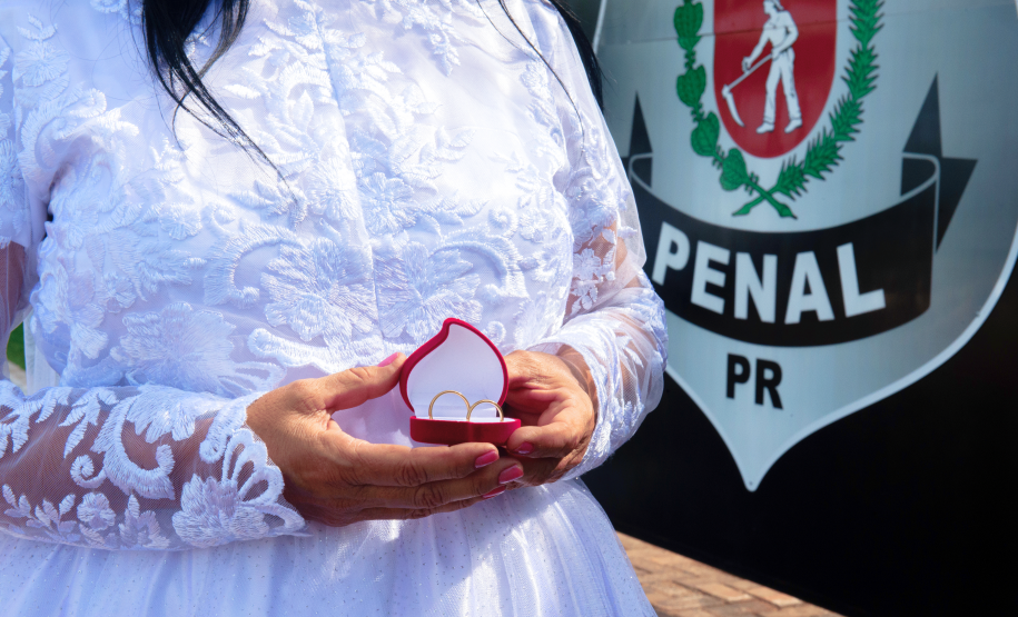 Imagem que mostra uma noiva com um vestido branco com aplicações de renda. Ela segura uma pequena caixa vermelha com duas alianças. Ao fundo é possível ver o brasão do DEPEN em uma parede.