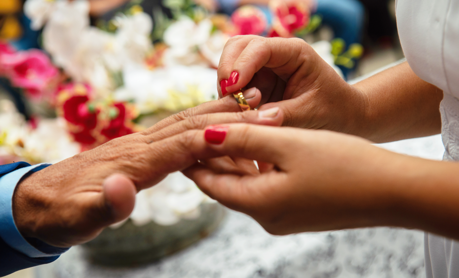 Imagem que mostra uma das noivas colocando a aliança no dedo do noivo. O fundo mostra flores desfocadas. É possível ver parte do vestido dela, branco. Ela tem as unhas pintadas com esmalte vermelho.