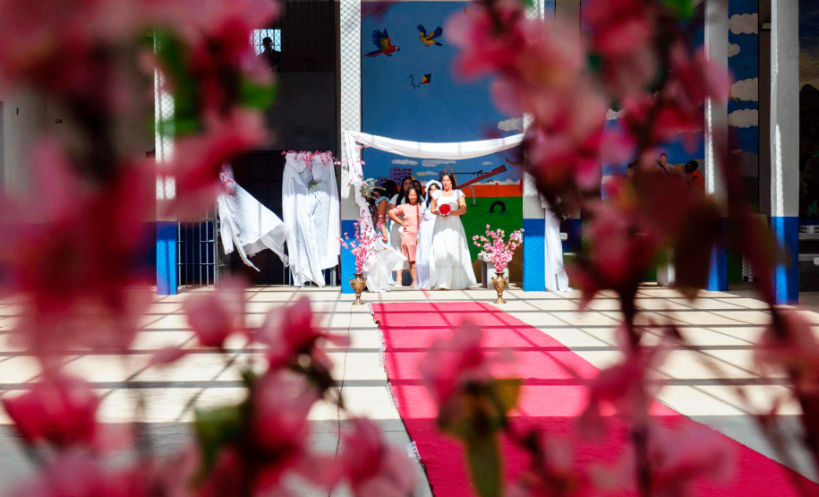 Imagem que mostra as noivas em fila preparadas para a entrada. O ambiente está decorado com panos brancos, flores rosas e um tapete vermelho.