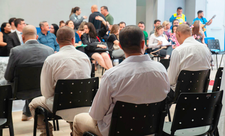 Imagem que mostra os noivos sentados em cadeiras pretas, de costas. Ao fundo é possível ver as autoridades presentes.