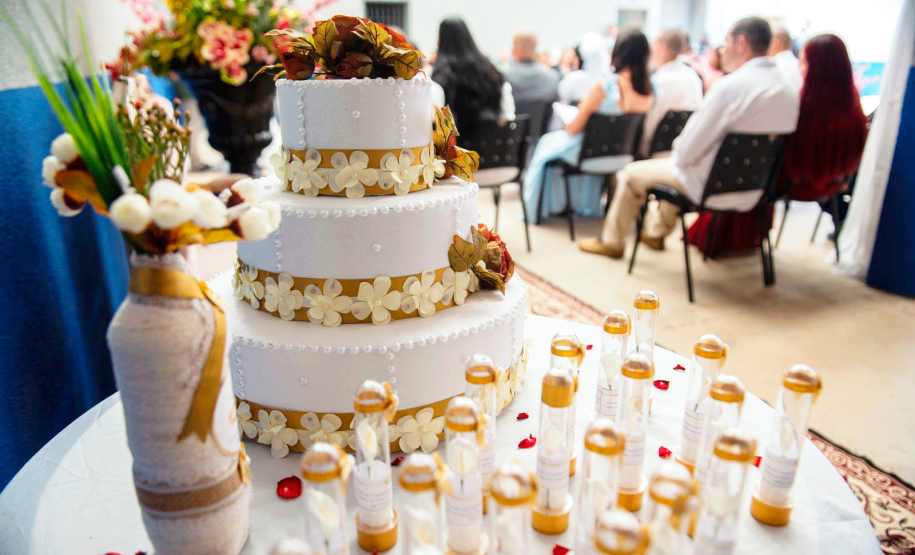 Imagem que mostra uma mesa decorada com um bolo, arranjo de flores e lembrancinhas em tons de branco e dourado. Ao fundo é possível ver os casais sentados, de costas.