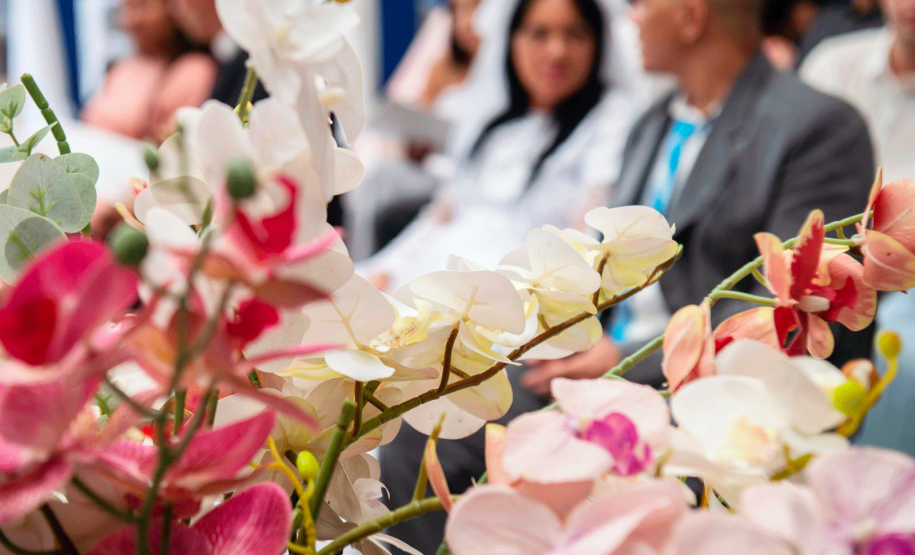 Imagem que mostra, em primeiro plano, flores de um dos arranjos da cerimônia. São orquídeas em tons de branco, rosa e vermelho. Ao fundo é possível ver um casal, de forma borrada.