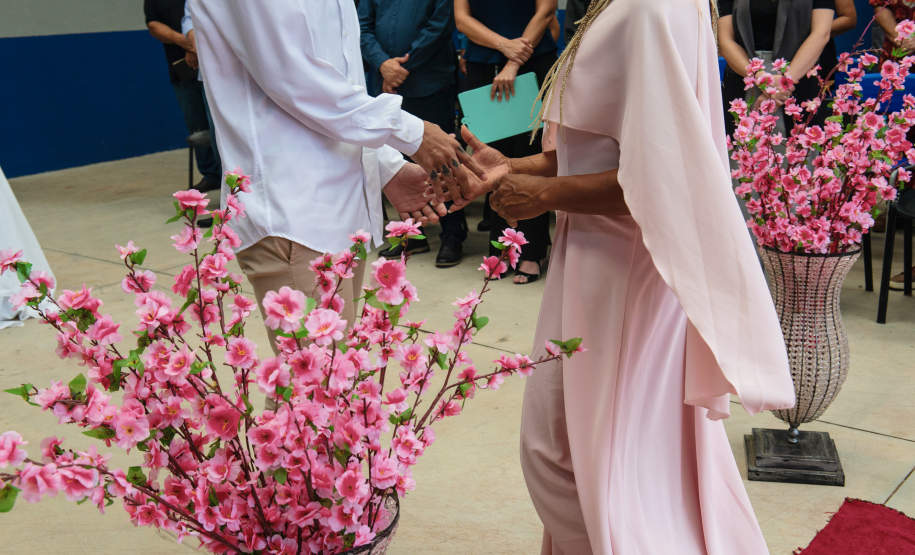 Imagem que mostra um dos casais, em pé, dando as mãos. Ela usa um vestido com capa em tom rosa claro. Ele usa uma camisa branca e calça bege. Ao fundo é possível ver as autoridades presentes assistindo a cena. Também há flores rosas em arranjos compondo a cena.