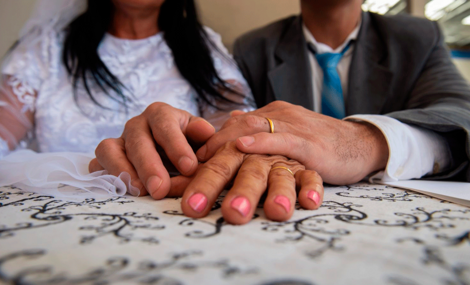casal com as mãos sobre a mesa mostrando as alianças. eles usam trajes de terno e gravata e vestido de noiva.