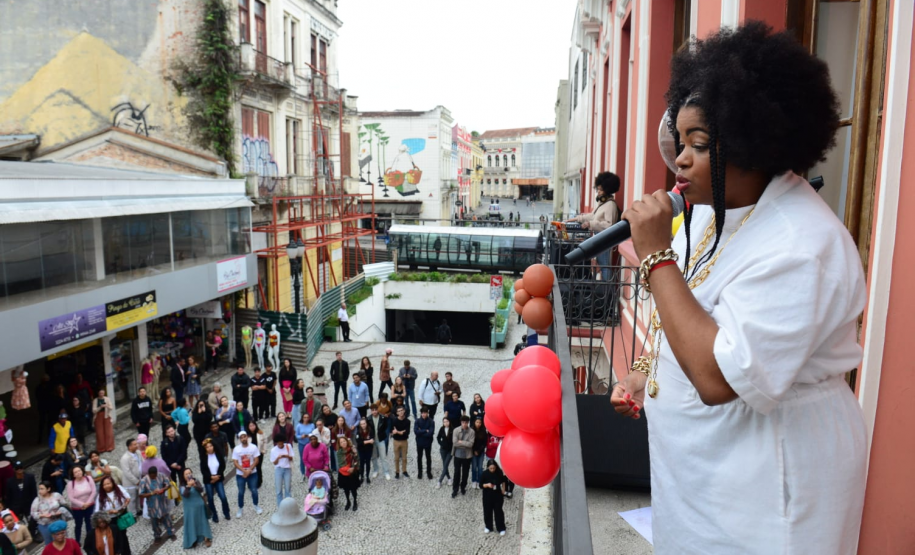 Imagem da cantora Janine Mathias fazendo sua apresentação na janela do prédio histório da sede central de atendimento da Defensoria, com o público ao fundo da imagem