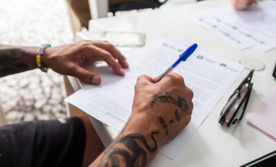 Imagem horizontal colorida em aparece o detalhe da mão de um usuário preenchendo documentos durante atendimento da DPE-PR.
