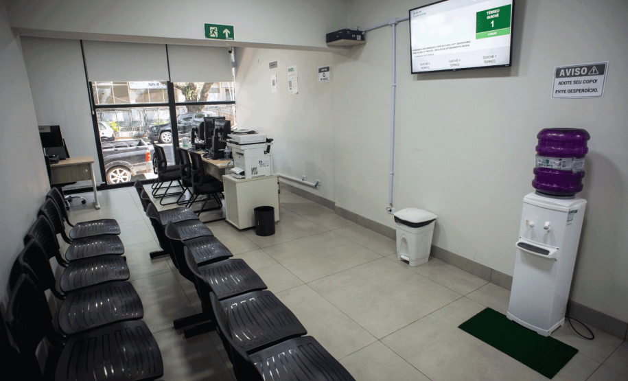 Espaço de espera com fileiras de cadeiras pretas para o público, um bebedouro de água, uma lixeira e um painel eletrônico na parede exibindo senhas de atendimento; no fundo, uma grande janela de vidro permite a visão da rua.