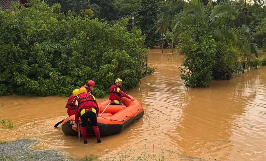 Imagem horizontal colorida em que se vê bombeiros em um bote inflável entrando em área alagada pelas enchentes no Litoral do Paraná.