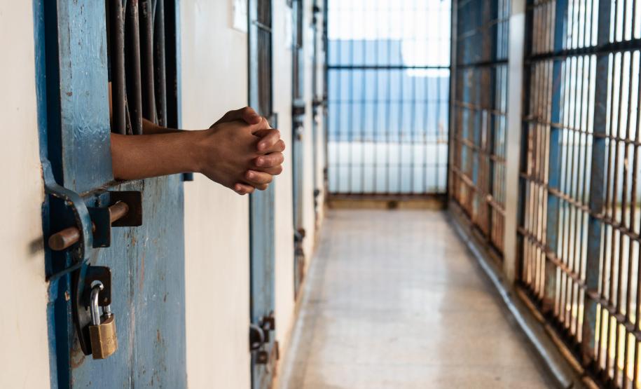 Imagem horizontal colorida em que se vê as mão se um jovem em primeiro plano e ao fundo o corredor da unidade socioeducativa.