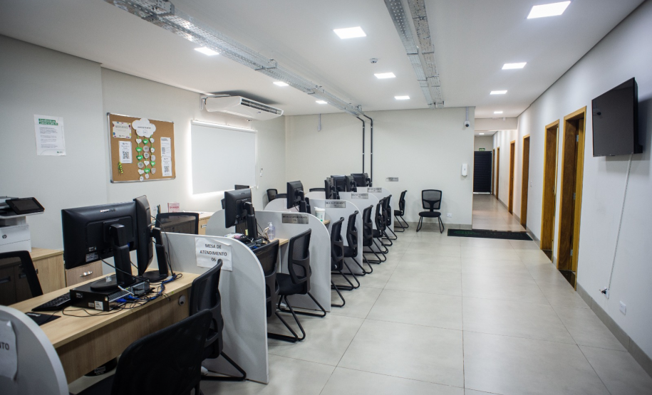 Sala ampla e bem iluminada com várias mesas de atendimento organizadas em fileiras, cada uma equipada com computador e cadeiras para os atendentes e assistidos; ao fundo, um corredor leva a outras salas.