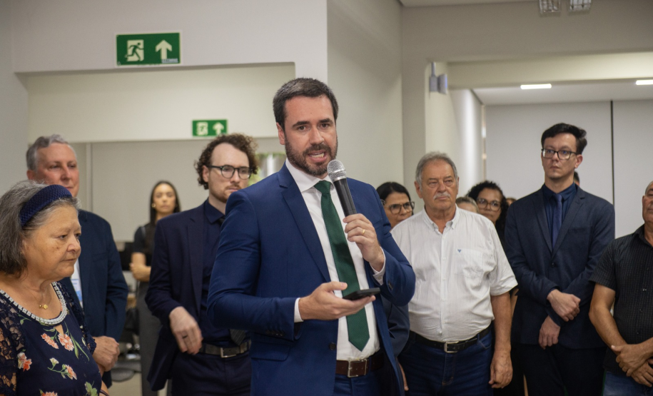 Defensor público-geral com um terno azul e gravata verde segura um microfone e um celular enquanto discursa para um público atento; ao seu redor, estão pessoas de diferentes idades e estilos, algumas vestindo roupas formais e outras mais casuais, em um ambiente fechado e iluminado.