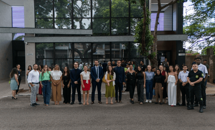 Autoridades e servidores da Defensoria Pública posam para foto na frente do prédio, vestindo trajes formais. Alguns seguram documentos e faixas comemorativas.