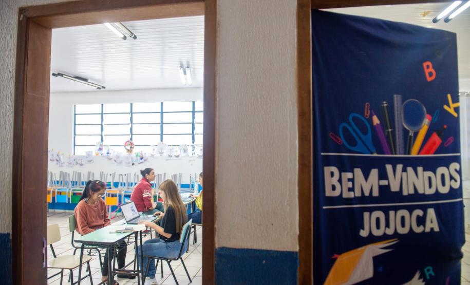 Imagem que mostra o interior de uma sala de aula vista através de uma porta entreaberta. Na sala, há pessoas sendo atendidas por agentes da DPE-PR. A sala possui janelas grandes com grades, permitindo a entrada de luz natural. Há decorações coloridas penduradas nas janelas e nas paredes. À direita, em primeiro plano, parte de uma faixa azul escura está visível, pendurada na porta, com a inscrição "BEM-VINDOS JOJOCA" em letras brancas grandes, acompanhada de desenhos coloridos de materiais escolares.