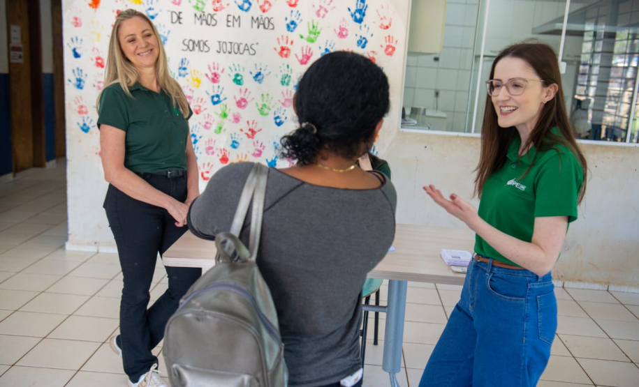 Imagem que mostra duas servidoras da DPE-PR interagindo com uma mulher. À esquerda, uma mulher loira, vestindo uma camiseta verde escura e calças pretas, sorri. Outra mulher está de costas para a câmera. Ela veste uma blusa cinza e tem uma mochila prateada. À direita, outra mulher com cabelos castanhos e óculos, vestindo uma camiseta verde e calças jeans azuis, gesticula com as mãos enquanto sorri. Ao fundo, na parede, há diversas impressões de mãos coloridas e a frase "DE MÃOS EM MÃOS SOMOS JOJOCAS".